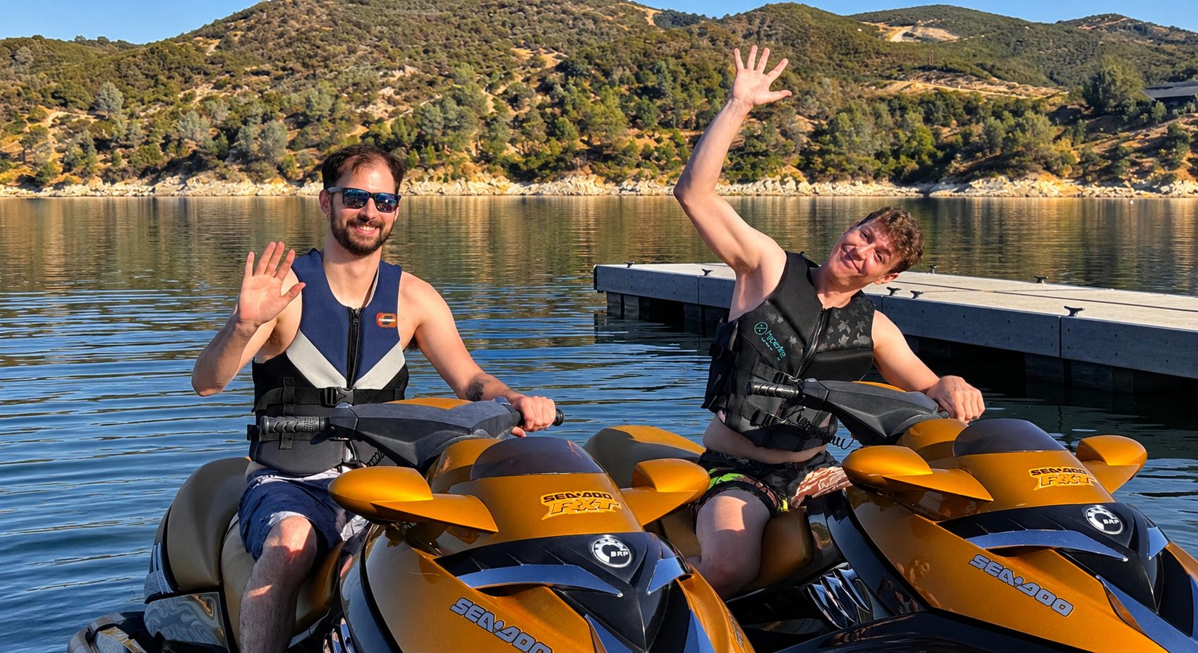 Two riders waving from jet skis on Lake Nacimiento, with oak-covered hills behind